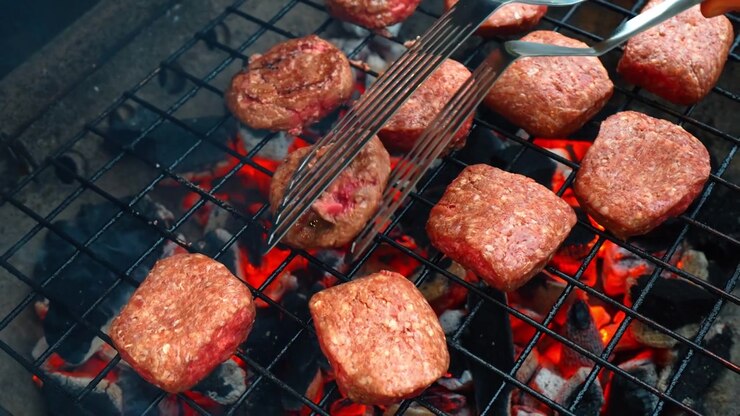 Metal spatula moving meat on hot charcoal grill. Point of view shot of metal spatula handling meat patties on barbecue grill with burning charcoal and smoke.