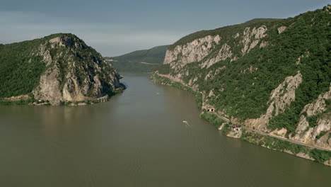 Impresionante-Toma-Aérea-De-Dubova,-Rumania,-Que-Captura-La-Serenidad-Del-Río-Que-Serpentea-A-Través-De-Majestuosos-Acantilados-Cubiertos-De-Verde-Y-Una-Carretera-Panorámica.