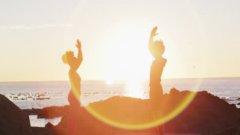 african american couple practicing yoga together on the rocks near the sea during sunset