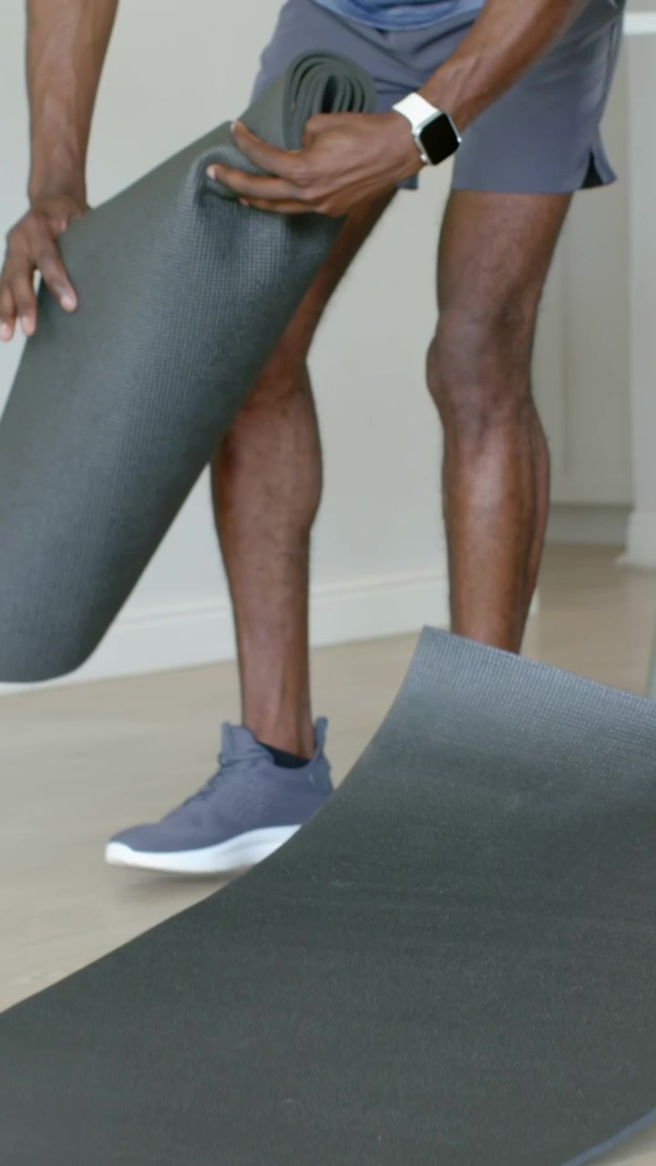 Vertical video: Closing eyes, man in blue tee adjusting and unrolling dark mat at home for workout