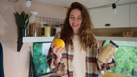 a happy brunette girl in a plaid shirt holds an orange and a jar of pasta in her hands and chooses something from them during her vacation in a trailer in a camp in the city in the summer