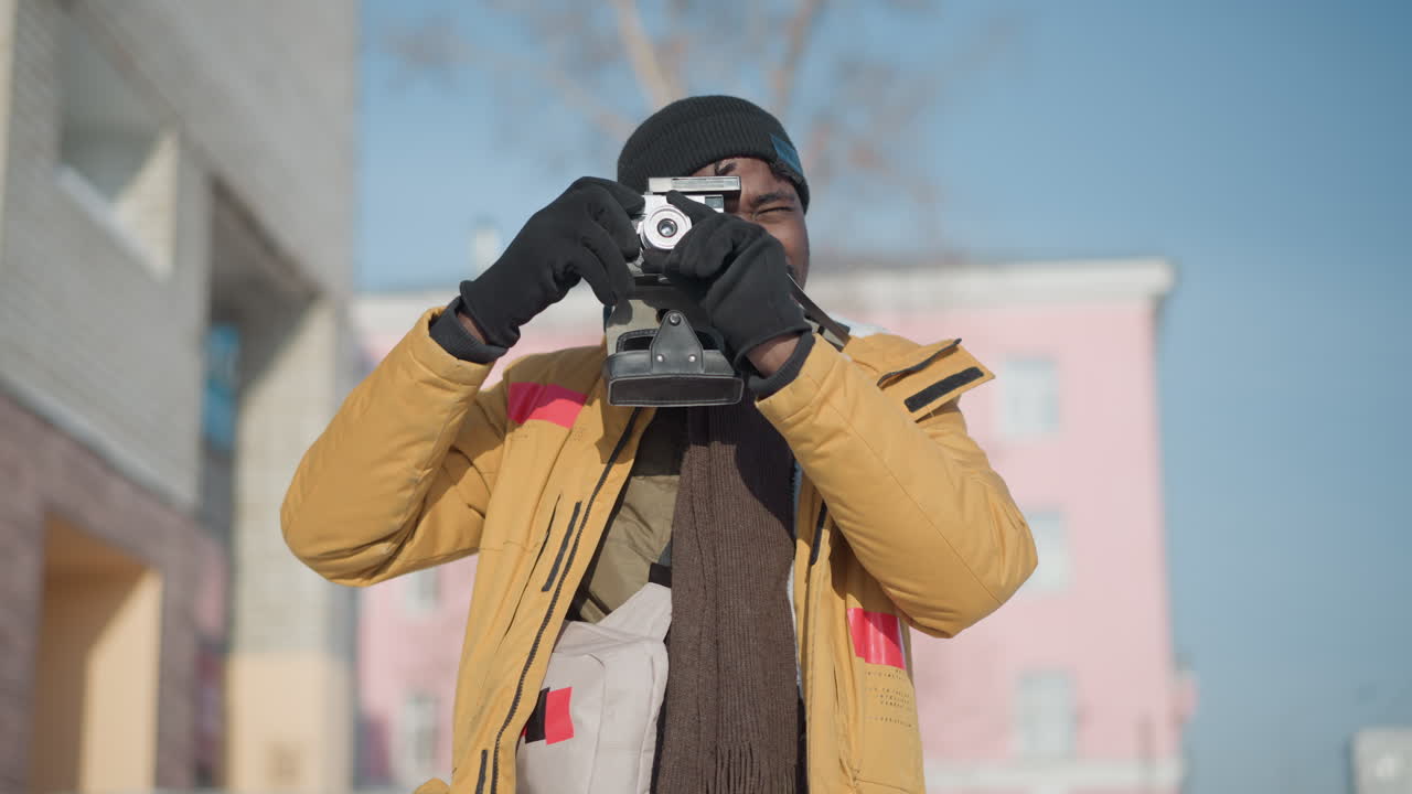young dark man carrying bag and camera fixes eyes on camera to take pictures walking snowy urban sidewalk under bright sun, blurred pedestrians and pink building in background, photography scene
