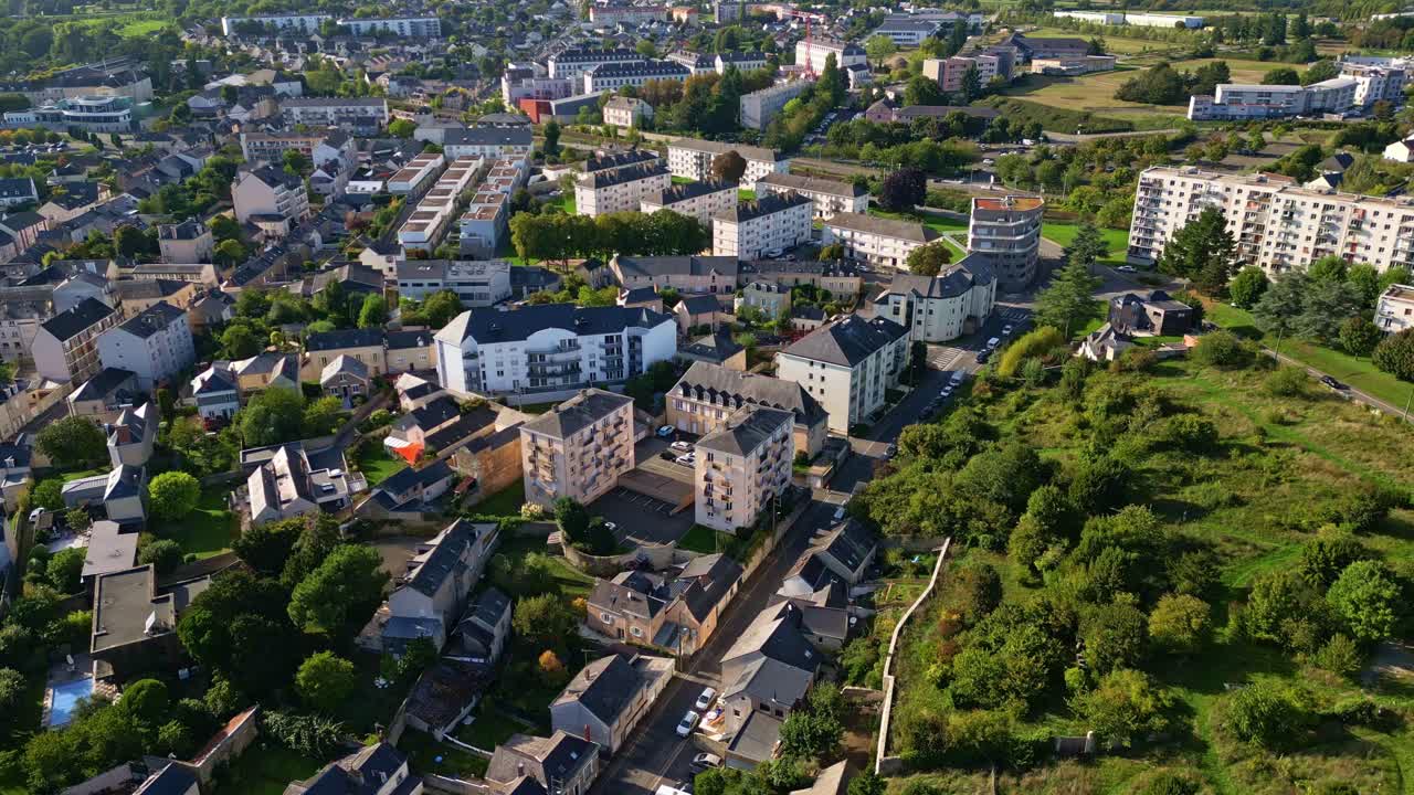 Slow drone glide over suburban Laval reveals contrasting urban forms with clusters of white apartment blocks, traditional houses, and the mosaic edge of green parks and open land, Brittany, France
