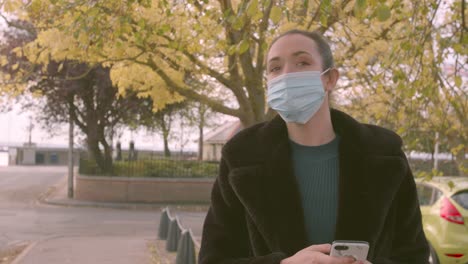 female wearing a medial facemask using phone and looking up for a bus