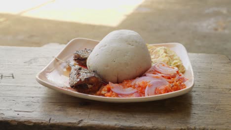 Una-Mujer-Pone-Sobre-La-Mesa-Un-Plato-Con-Banku,-Una-Típica-Bola-De-Masa-Ghanesa,-Acompañada-De-Pescado-Y-Otras-Guarniciones,-Almuerzo-Ghanés