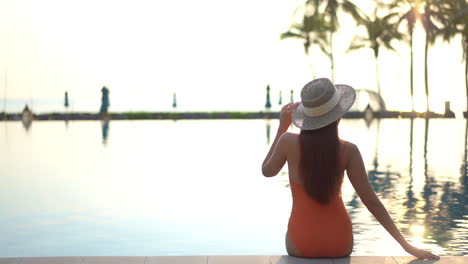 Vista-Trasera-De-Una-Mujer-Sentada-En-El-Borde-De-La-Piscina-Al-Atardecer-Con-Traje-De-Baño-Naranja-Y-Sosteniendo-Un-Sombrero-Ancho