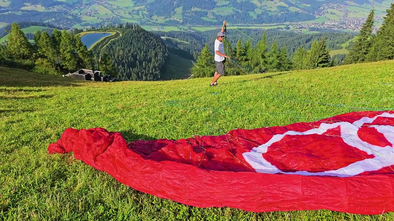 Avusturya, Kitzbühel'de yamaç paraşütü kurulumu; yeşil bir alp çayırına serilmiş kırmızı paraşüt, ipleri hazırlayan pilot, vadi manzaraları, çam ormanları ve yazın engebeli Tirol dağları