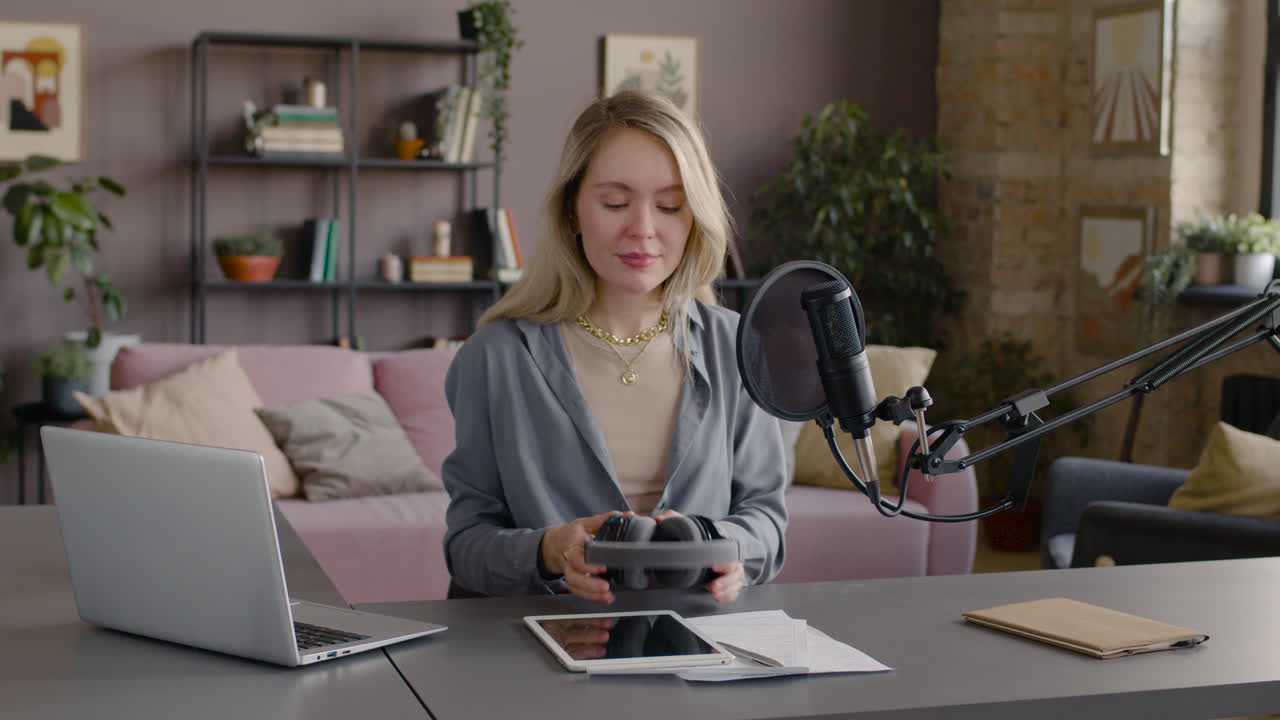 Woman Putting On Headphones While Talking Into A Microphone Sitting At A Desk With Laptop, Tablet And Documents