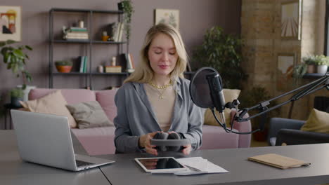 woman putting on headphones while talking into a microphone sitting at a desk with laptop, tablet and documents