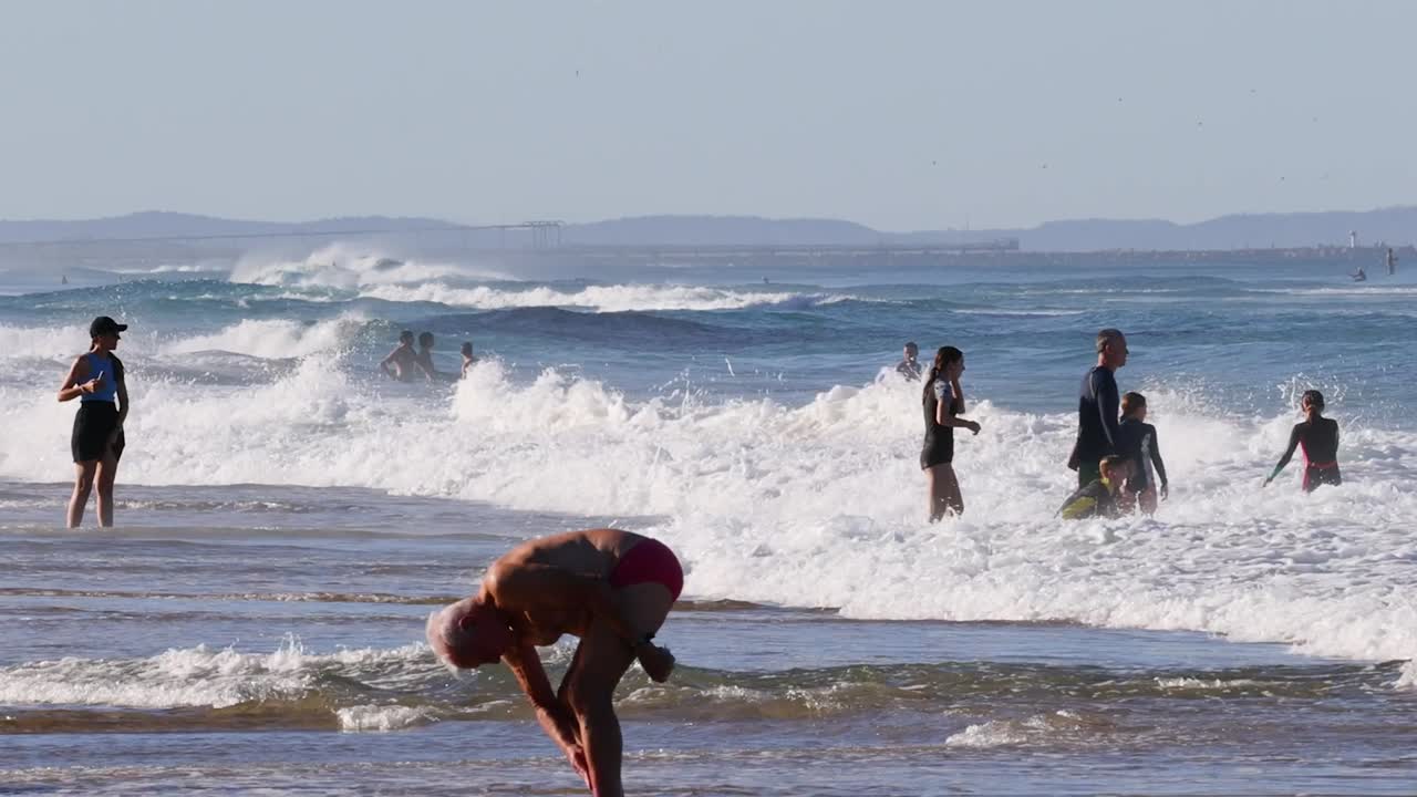 People enjoying the surf and waves, with surfers carrying boards and beachgoers playing in the water.