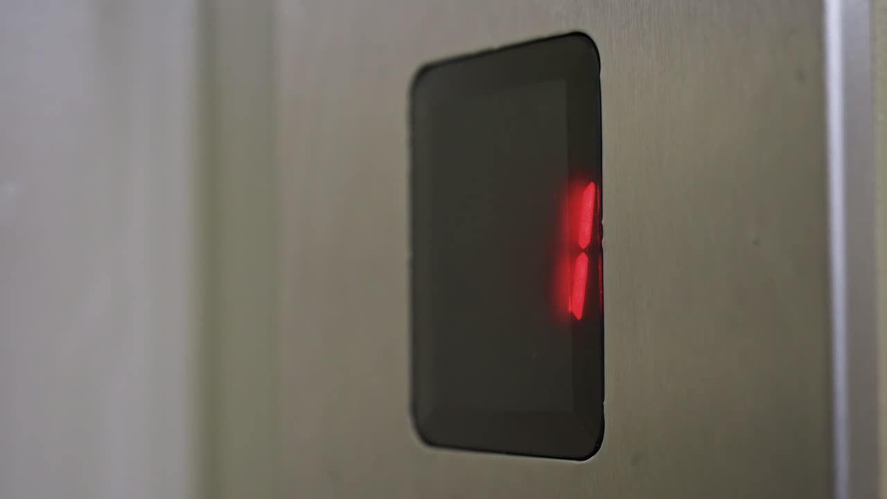 Primo piano della luce rossa dell'indicatore di piano dell'ascensore che si illumina, cifra numerica illuminata su pannello in acciaio inossidabile all'interno di un corridoio fioco, movimento sottile che suggerisce l'arrivo dell'ascensore, atmosfera interna urbana minimale, fredda.
