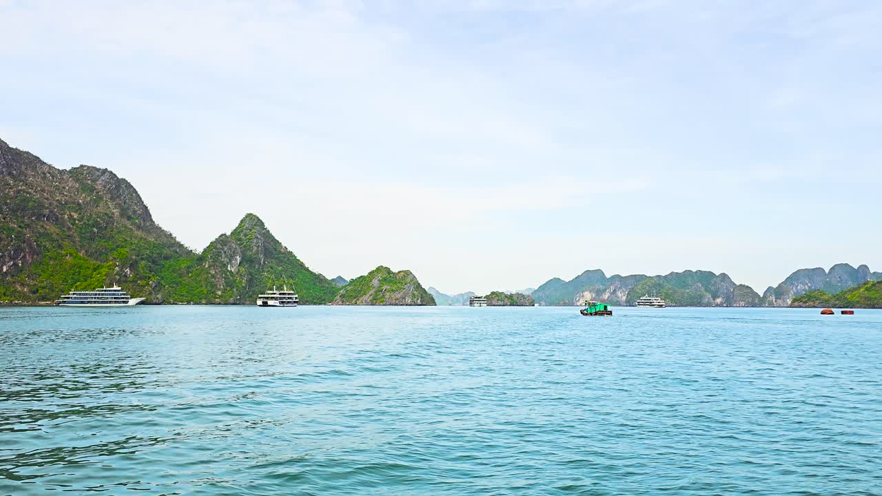 Emerald waters of Ha Long Bay stretch between dramatic limestone karsts rising from the sea, while tourist boats and luxury cruise ships navigate through the tranquil seascape under a soft blue sky