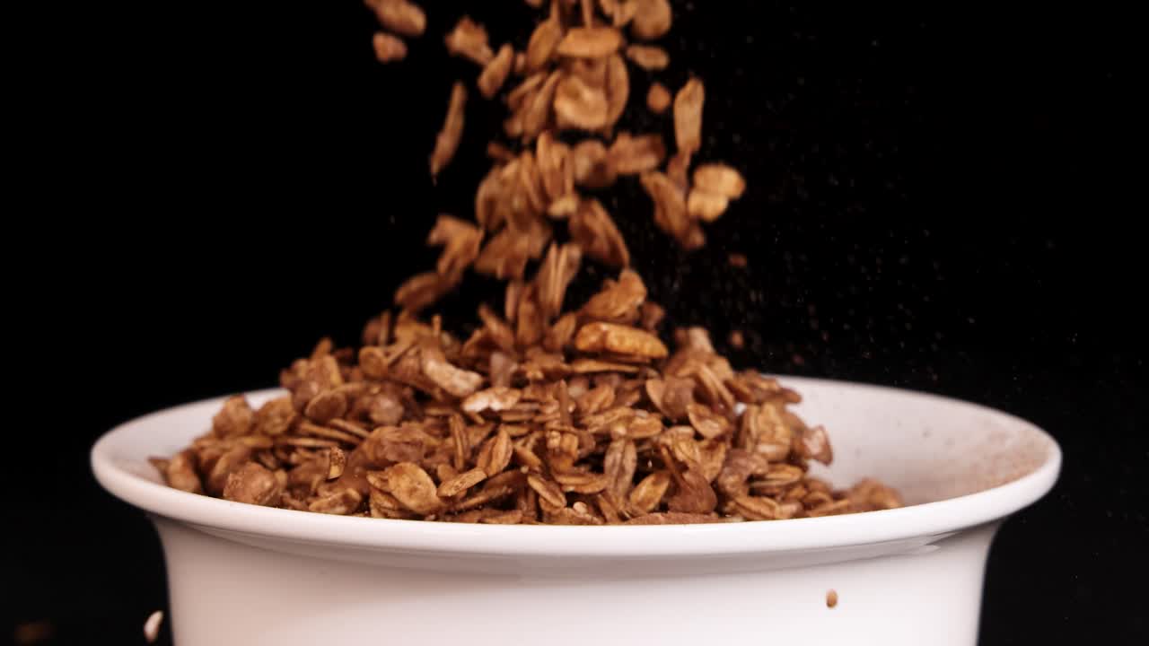 Granola grains cascade into a white bowl, set against a black background, highlighting texture and movement in slow motion