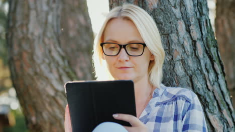 attractive young woman in glasses uses a tablet sits in a park near a tree beautiful light before su