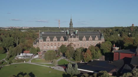 Wide-aerial-view-of-Nordiska-Museet-with-Kaknästornet-and-forest-in-background-on-sunny-evening-on-Djurgården-in-Stockholm,-Sweden