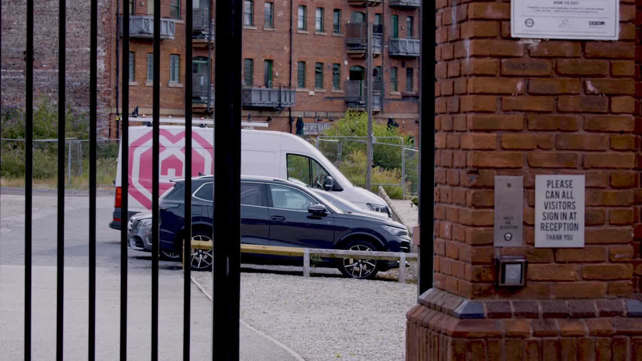 En sikkerhedsport åbner og afslører en parkeret bil og en varevogn på en moderne byparkeringsplads, med stabilt dagslys og statisk kameraføring.