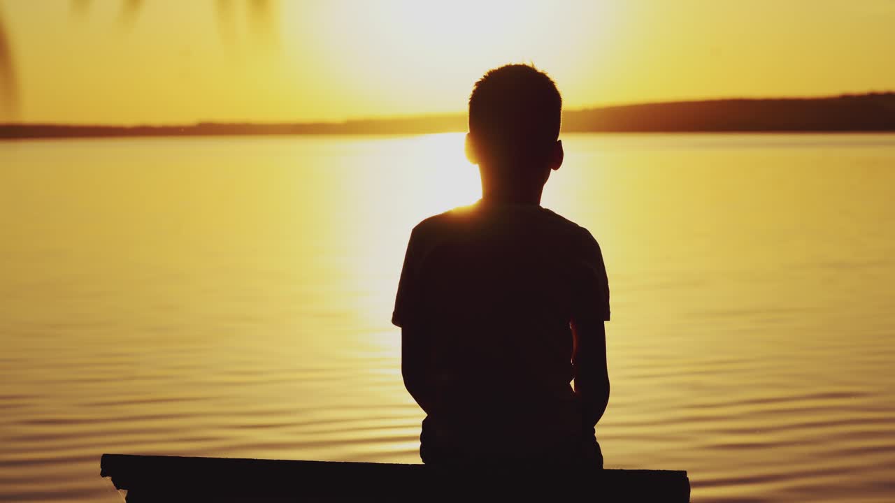 Little boy sitting on wooden dock at sunset. Child is watching the sunset against the background of a river. Dreams Come True.
