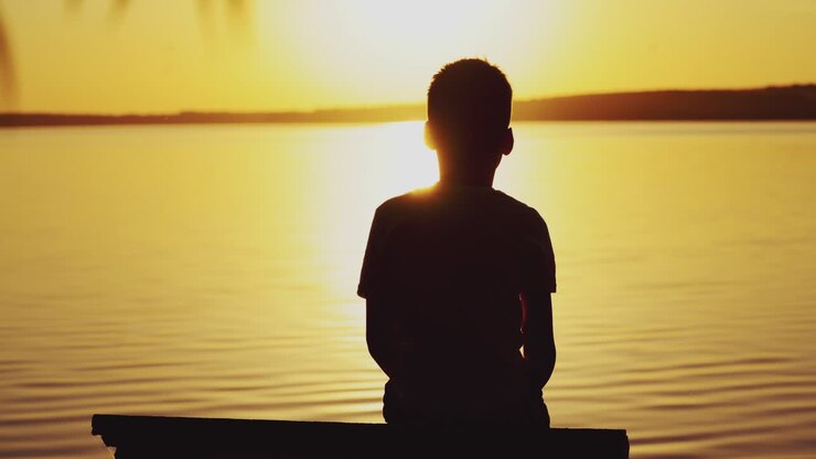 Un bambino seduto su un molo di legno al tramonto. Il bambino guarda il tramonto sullo sfondo di un fiume. I sogni diventano realtà.