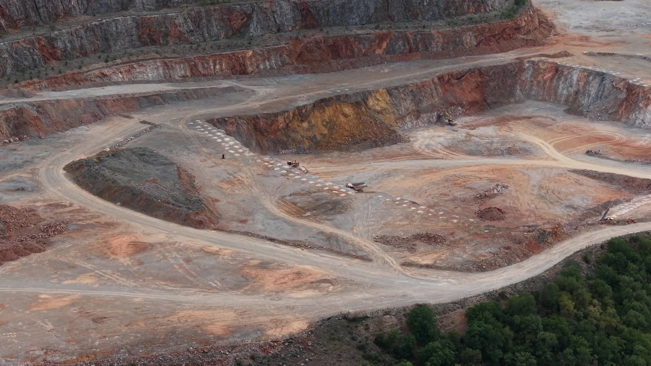 An aerial wide view of an active open pit mine site showing red earth, tiered rock levels, and heavy machinery.