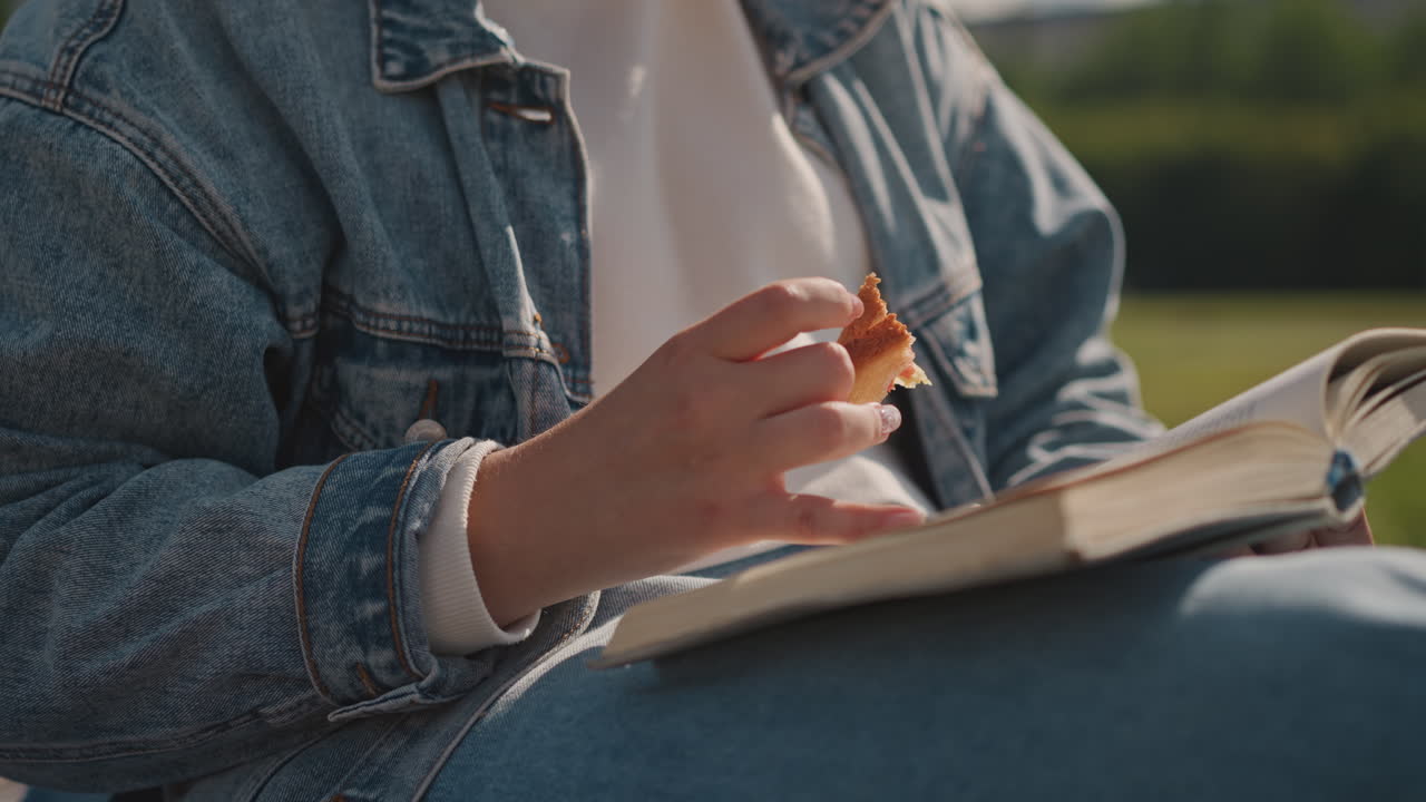 Kaukasisk student tar notater utendørs i dongerijakke med spiralblokk på fanget, penn holdt mellom fingrene, snack-øyeblikk fanget, fokusert studieenergi på solrik campusplen, produktiv livsstil.