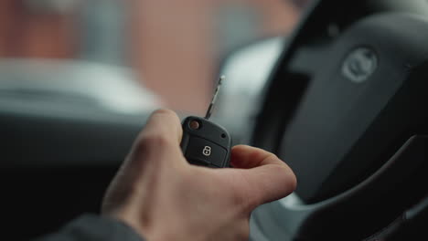 close up of male hand holding car key fob with lock and unlock buttons inside vehicle cabin focusing on ignition readiness and blurred steering wheel and dashboard with winter jacket sleeve visible