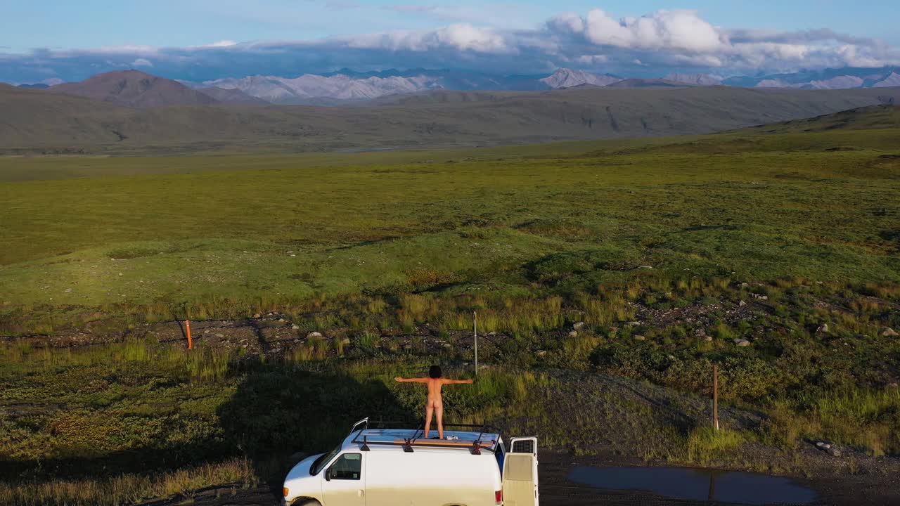 vista posterior de un hombre hipster desnudo parado en el techo de una furgoneta con los brazos abiertos estirados hacia las montañas de alaska brooks y campos verdes en verano