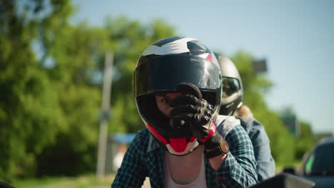 two friends ride a red power bike, both wearing helmets, with the rider closing her visor, the rider wears black gloves and a wristwatch, a car is visible behind them