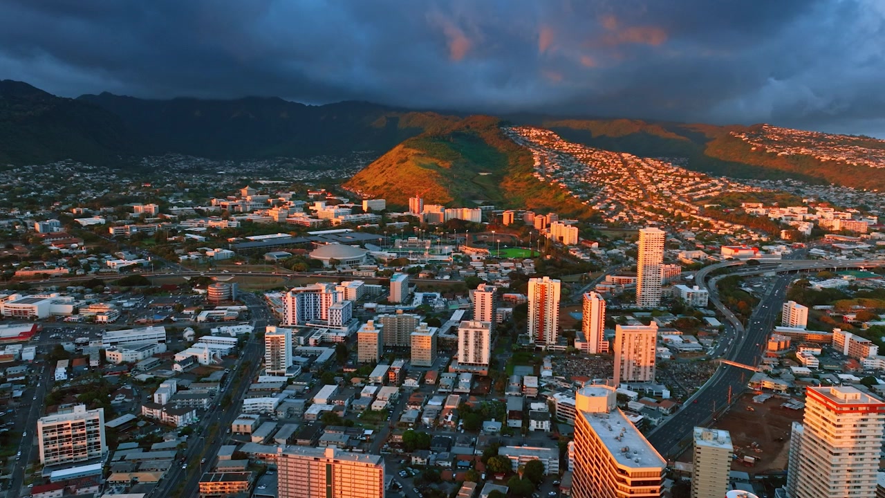 Riprese sopra il territorio urbano di Honolulu, Hawaii, USA al tramonto. Un drammatico cielo nuvoloso scuro copre il cielo sopra le montagne sullo sfondo