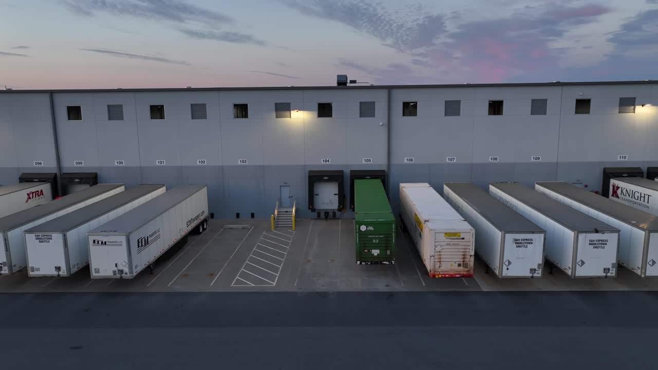 Formation of parked semi-trailers on ramp of warehouse at dusk. American factory building in Pennsylvania, USA. Aerial lateral wide shot.
