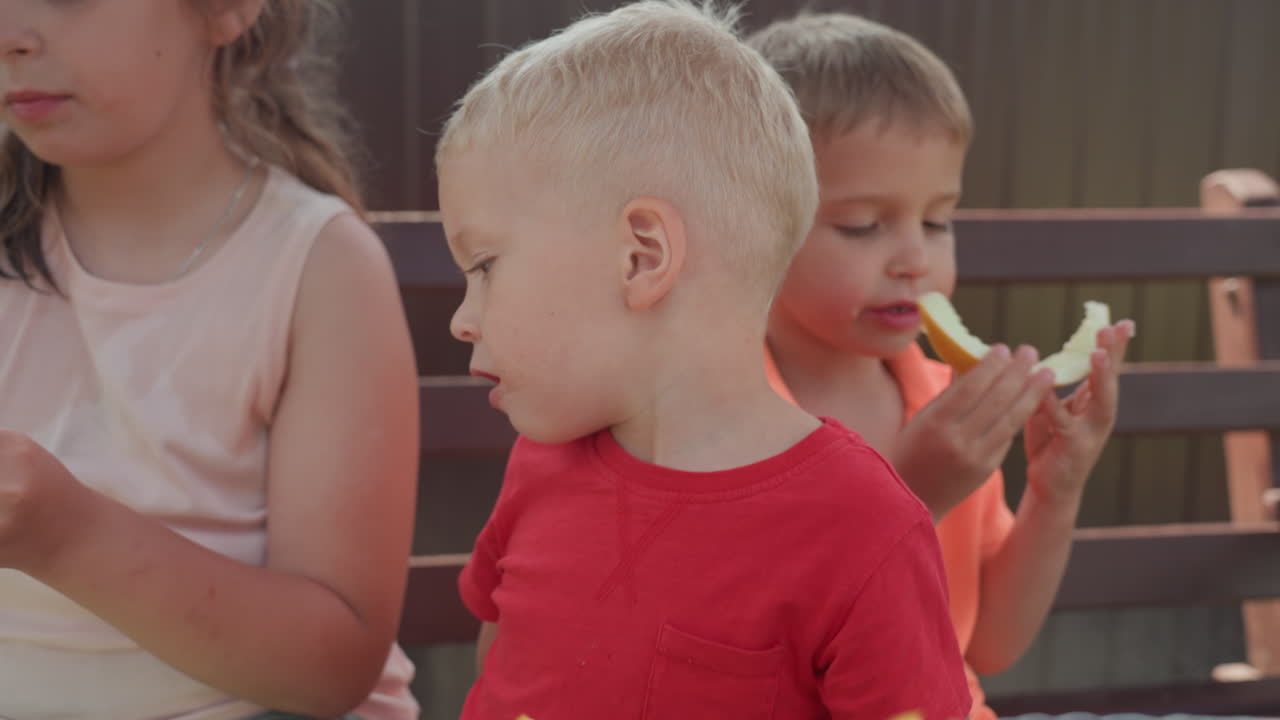 Youngsters Happily Eat Sweet Produce, Juvenile Kids Enthusiastically Savor Juicy Edible Portions Outdoors, Young Children Joyfully Share And Indulge In Luscious Fruit Slices Outside
