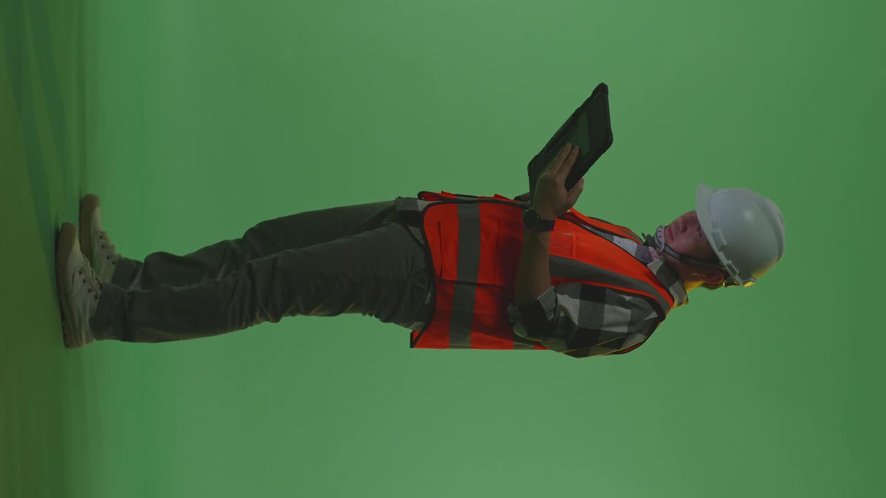 vista lateral completa del cuerpo de un ingeniero masculino asiático con casco de seguridad mirando la tableta en su mano y mirando a su alrededor mientras está de pie en el estudio de fondo de pantalla verde, durante la puesta o el amanecer