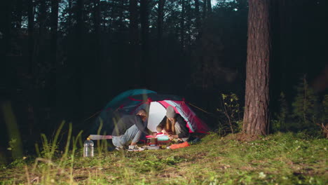 young couple prepares food together beside lit tent during evening forest camping, surrounded by trees and grassy ground, focused on cooking over portable stove