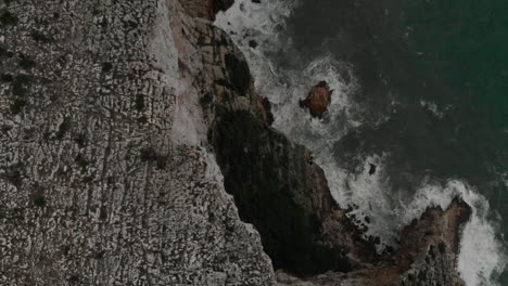 4k aerial view of a rock cliff with waves and green water sea in denia, alicante, spain, wide angle backward shot