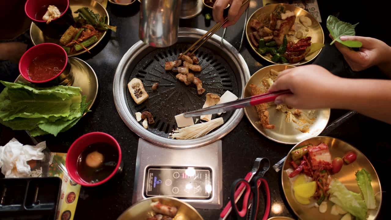 Hands grilling pork belly on tabletop grill, surrounded by side dishes, lettuce, and sauces