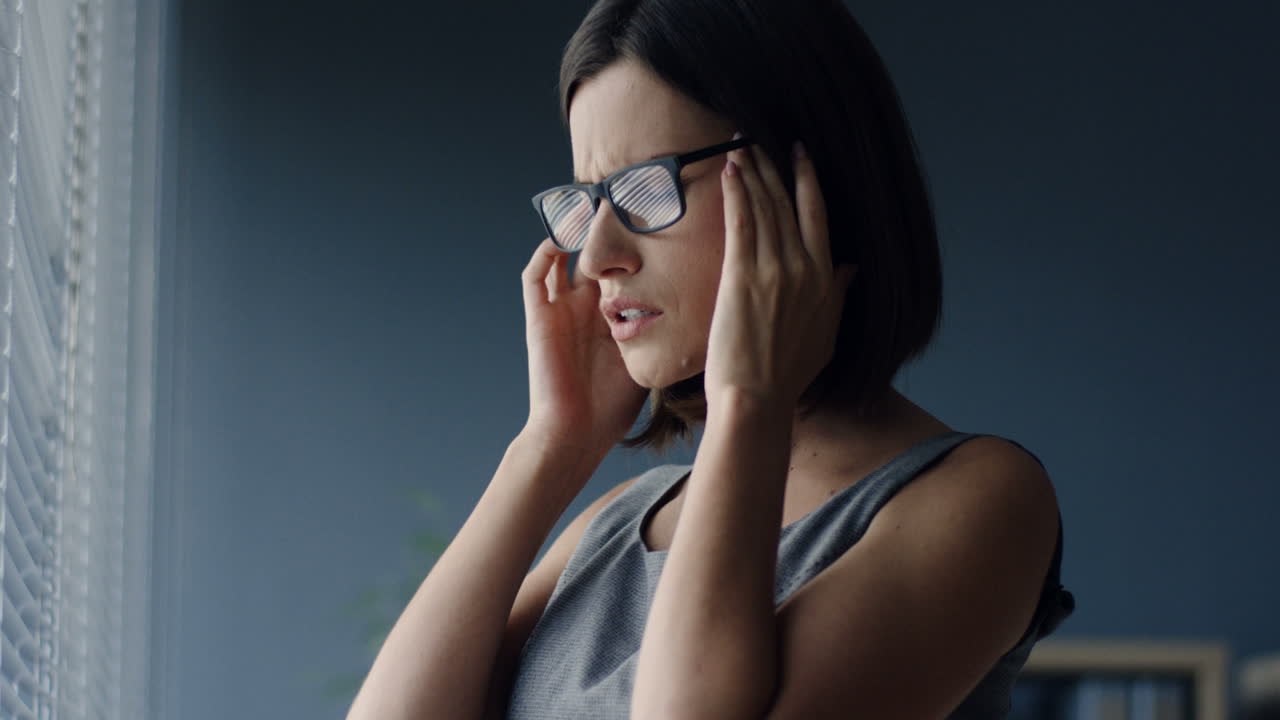 vista de cerca de una joven mujer de negocios con anteojos parada en la ventana de la oficina y con un fuerte dolor de cabeza