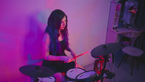 woman playing electronic drums in home studio with serious expression, surrounded by glowing pink and purple ambient light, focused on performance while seated near shelf and stool in creative setup