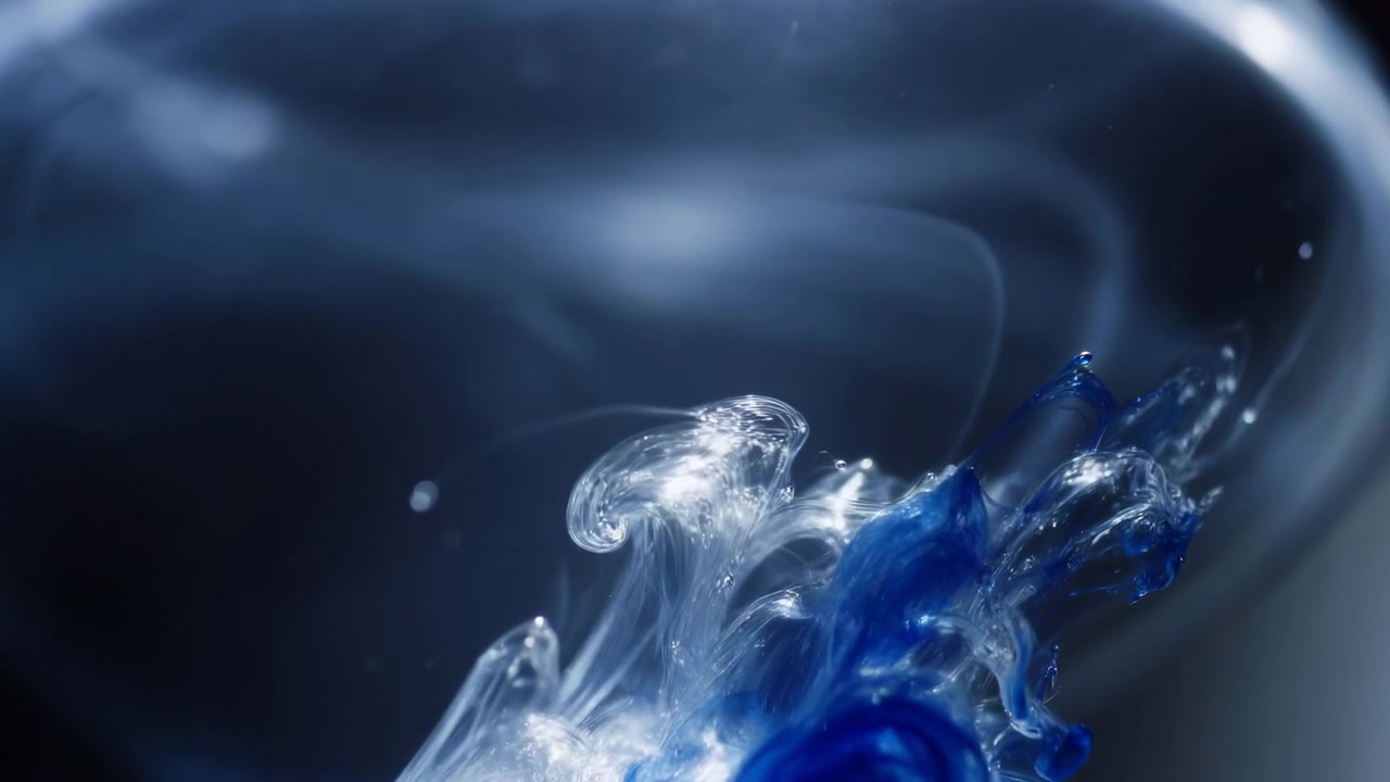 Low-angle close-up shows vivid blue pigment released into clear water, forming glossy folds, suspended droplets, thin filaments, microbubbles, and luminous contrast against deep navy studio background