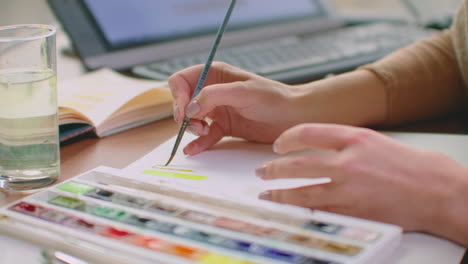 a young woman with a brush and paints chooses a suitable shade for the logo design or walls. choice of color and shade for future design. a woman colorist in an office with large windows draws with a brush and paints in a notebook.