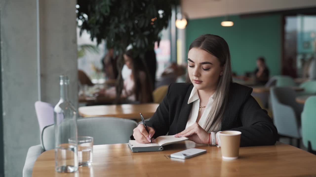 una giovane donna elegante si siede in un caffè in abito da lavoro, scrive in un quaderno con una penna, sorridendo