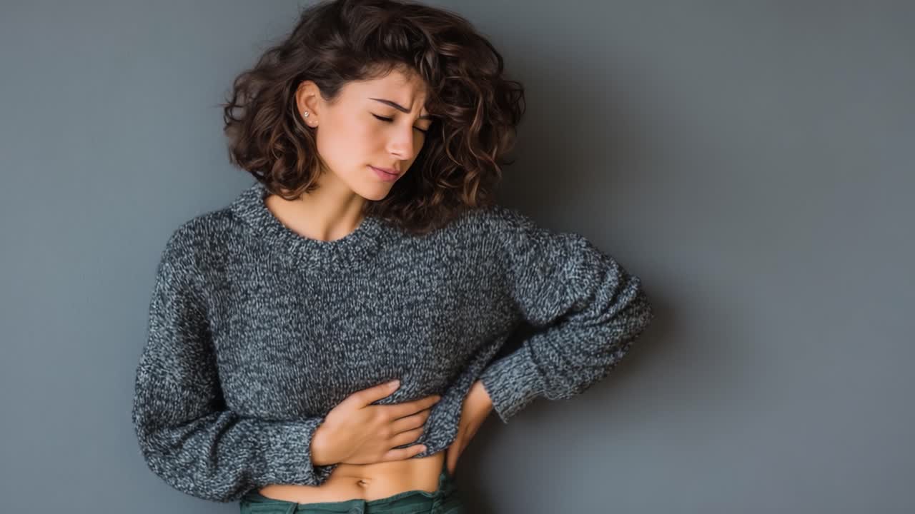 Una giovane donna con i capelli ricci che si tiene lo stomaco per il disagio, su uno sfondo grigio neutro, illustrando un momento di angoscia e malessere.