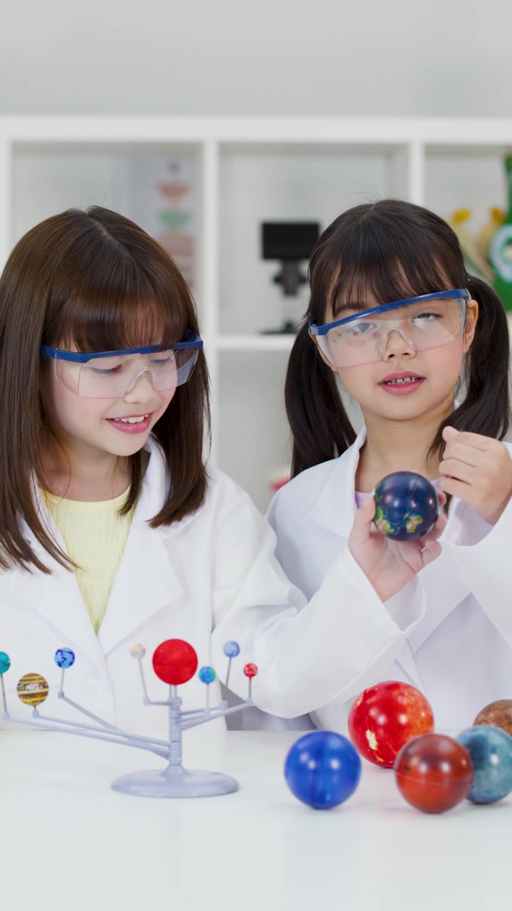 Two girls wearing safety goggles and lab coats examine a planetary model in science classroom.