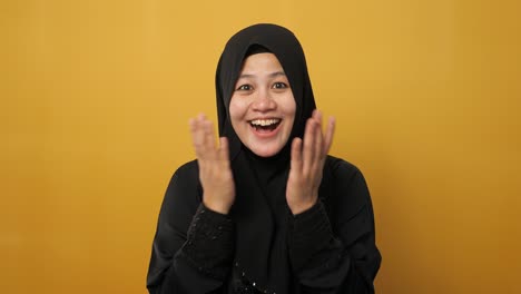 portrait of cute young asian muslim lady wearing hijab shows surprised or shocked expression with big eyes and open mouth, against yellow