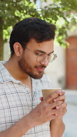 Happy-Indian-young-man-enjoying-morning-coffee-hot-drink-and-smiling-standing-on-city-street