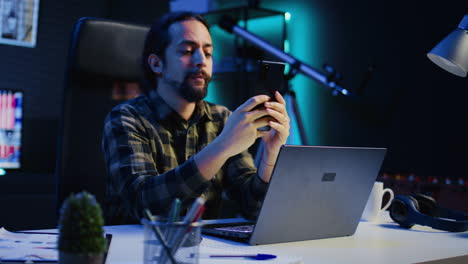 man relaxing at home sitting at office desk, texting friends on smartphone
