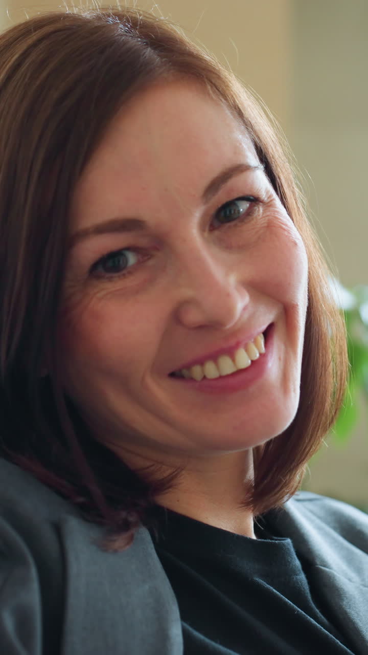 Smiling businesswoman in gray suit sitting casually on couch in cozy modern office with indoor plants and sunlight, projecting warmth, confidence, and approachability in corporate environment