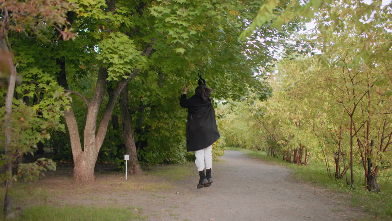 스타일리쉬한 관광객이 공원 산책로를 따라 낮은 가지에서 가을 잎을 따기 위해 점프하고, 녹색 캐노피 아래에서 공중으로 손을 뻗어 조용한 숲에서 장난기 넘치는 순간을 보내고 있어요. 부드러운 빛, 계절의 색, 캐주얼한 코트가 돋보이네요.