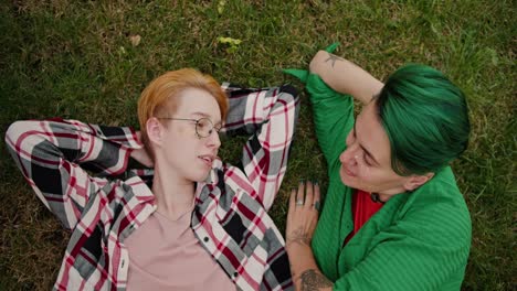 close-up shot of a blonde girl with a short haircut in glasses with sparkles on her face in a checkered shirt and her girlfriend with a short green haircut in a green shirt are talking lesha on the green grass in the park