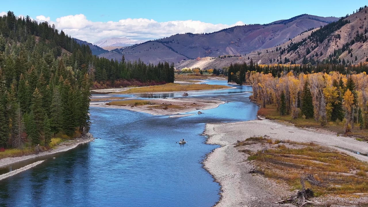 River bend with sandy shoreline and autumn forest in Wyoming valley, fisherman on kayak floats slowly, aerial telephoto tracking right