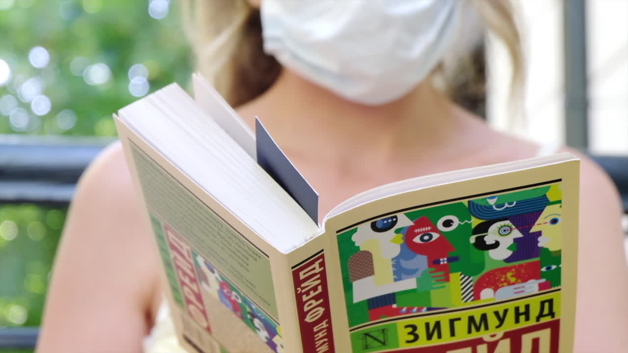 Chisinau, Moldova - August 18, 2021: Woman wearing a mask, reading Group Psychology and the Analysis of the Ego book by Sigmund Freud in Russian, at a terrace