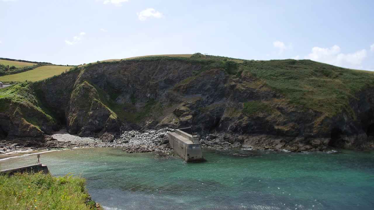 A breakwater is extending into calm water below steep cliffs on the Cornwall coast, located near Port Isaac and representing Portwenn with Doc Martin